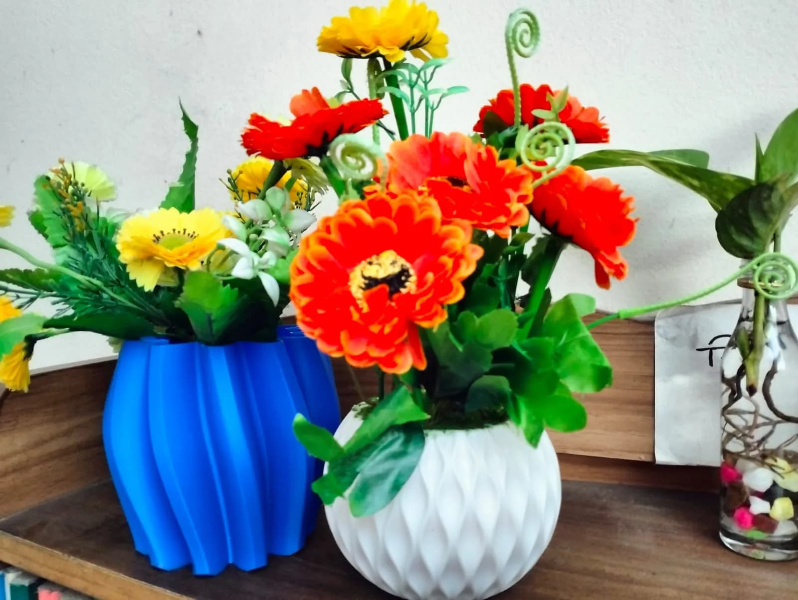 The blue twisted vase holding artificial flowers on a wooden shelf next to a white faceted pot