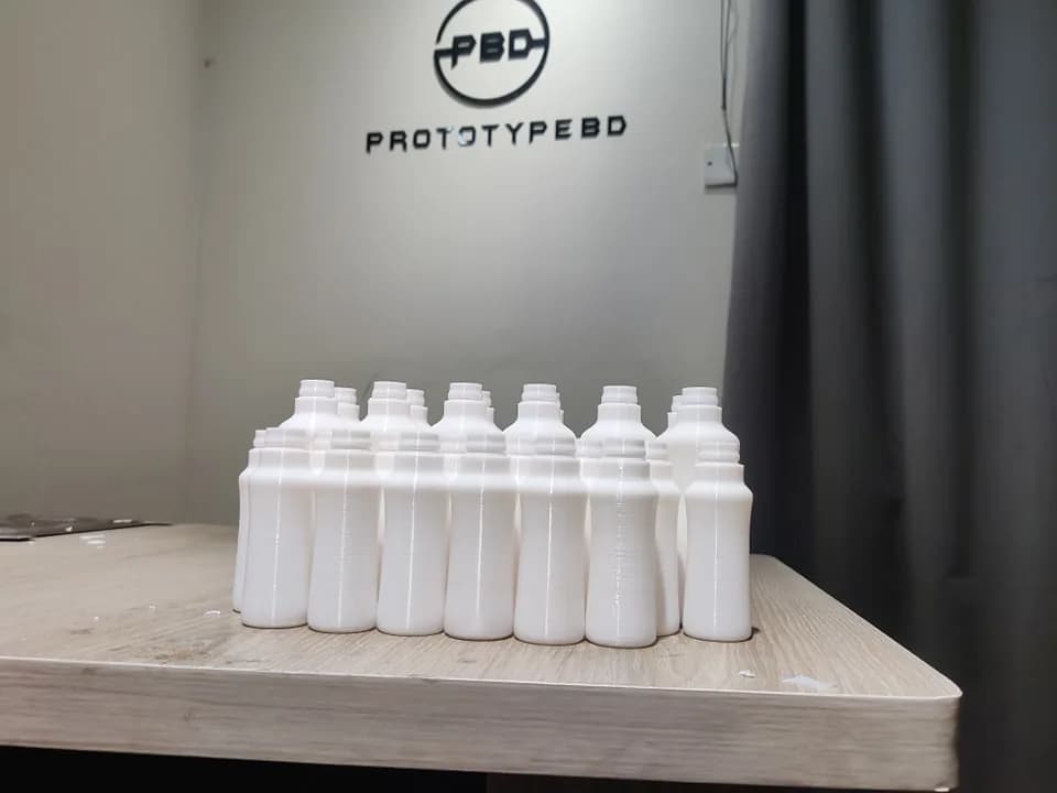 Batch of white bottle prototypes standing on a table in front of the PrototypeBD logo on the wall