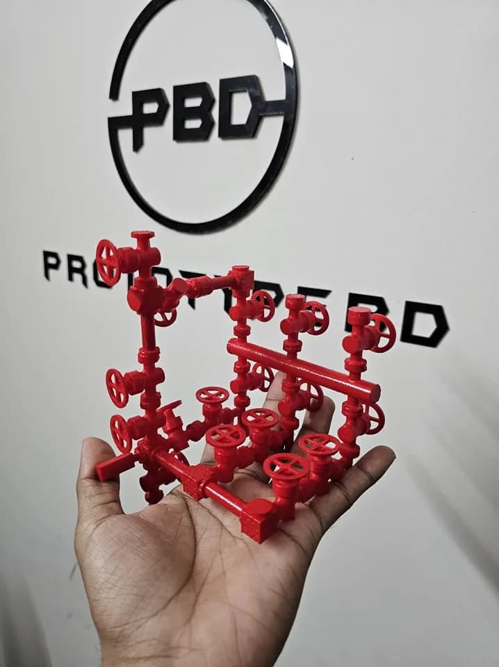 Red printed choke manifold model held up in front of the PrototypeBD logo on the workshop wall