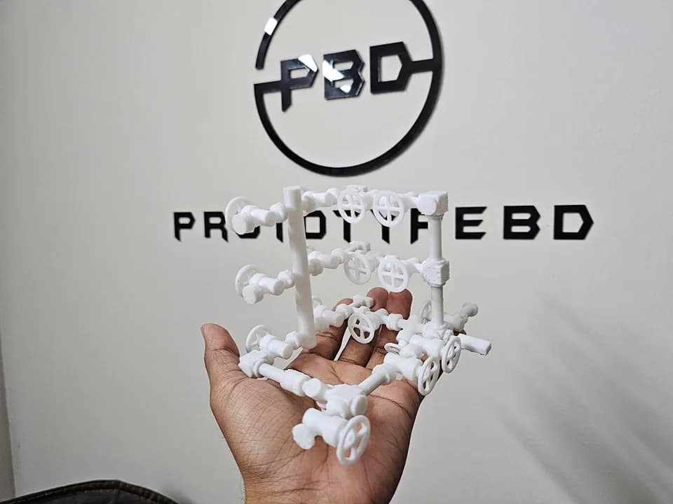 White version of the 3D printed choke manifold model held in front of the PrototypeBD wall sign