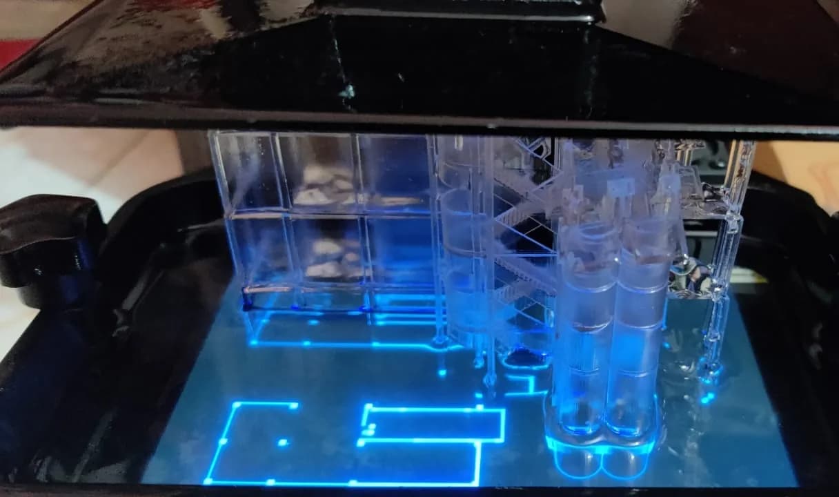 Clear resin building part hanging from the build plate inside an SLA printer, blue-lit layer outline visible below