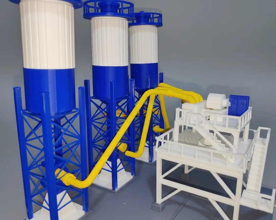 Close-up of the batching plant model's cement silos on lattice legs and the yellow conveyor tubes to the mixer platform