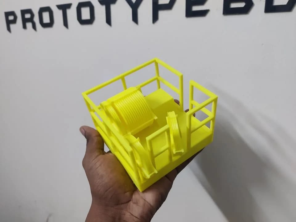 Hand holding the yellow printed crown block model in front of the PrototypeBD wall sign, showing the drum and grooved sheaves inside the railing