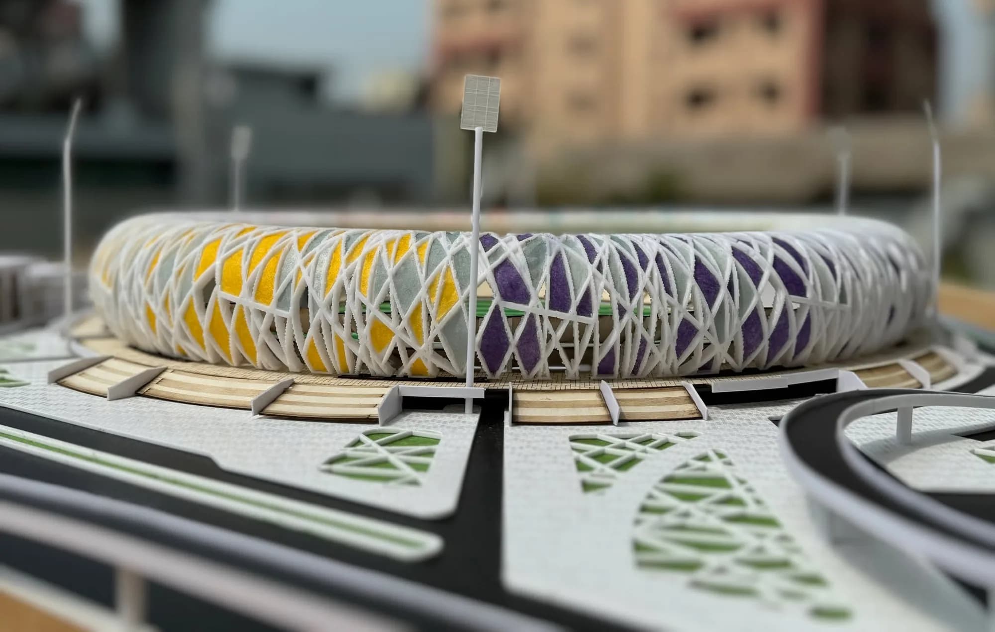 Low view of the stadium model's white lattice shell with yellow and purple panels and a floodlight mast