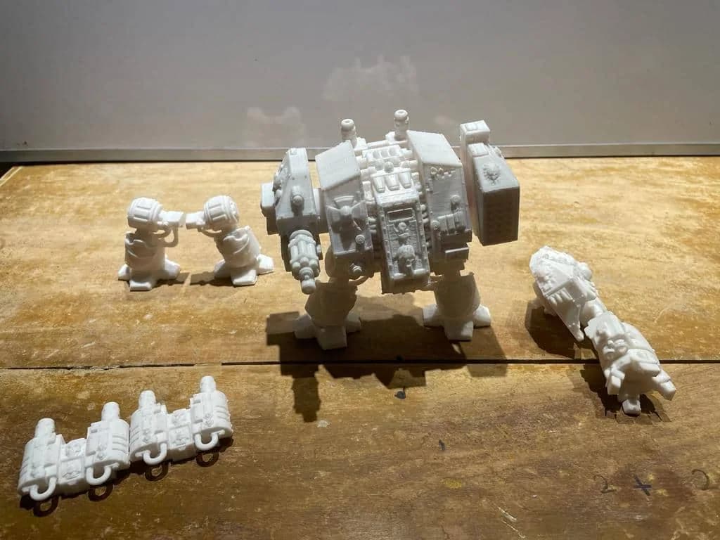 White 3D printed Dreadnought figure with spare weapon arms and accessory parts laid out on a wooden table