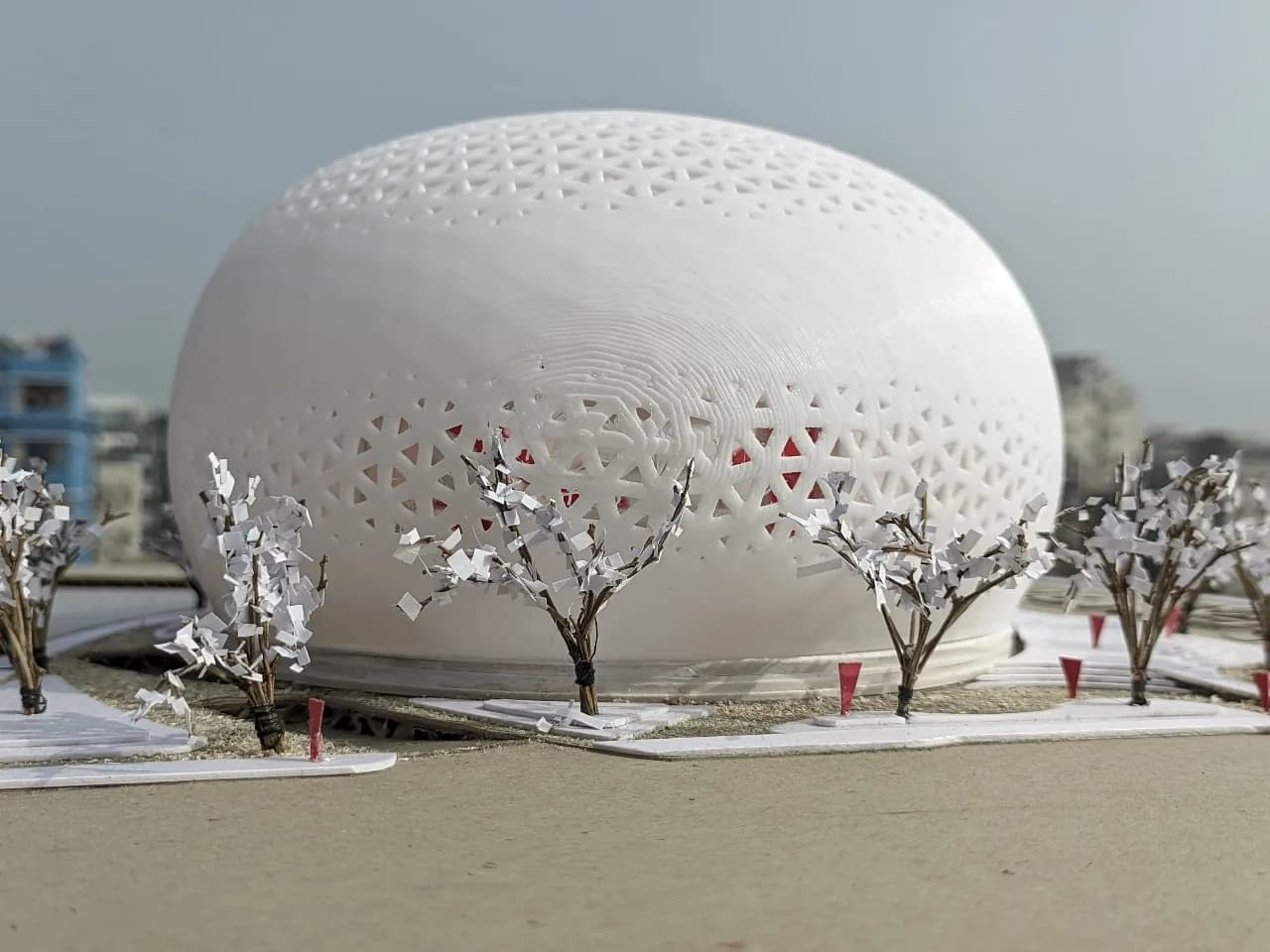 Eye-level view of the perforated exhibition hall shell with red interior showing through, behind small model trees