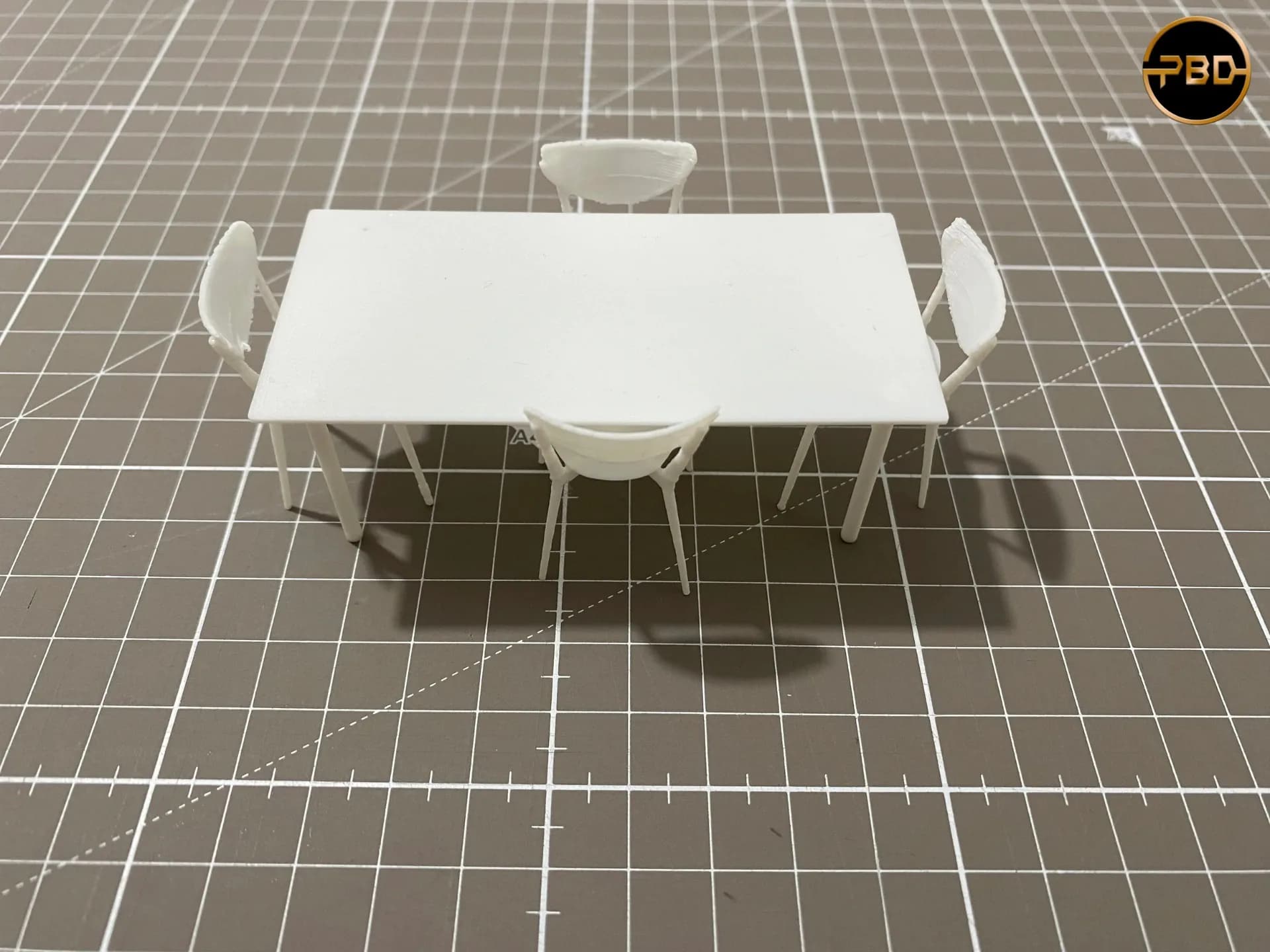 3D printed miniature dining table with four thin-legged chairs on a gridded cutting mat