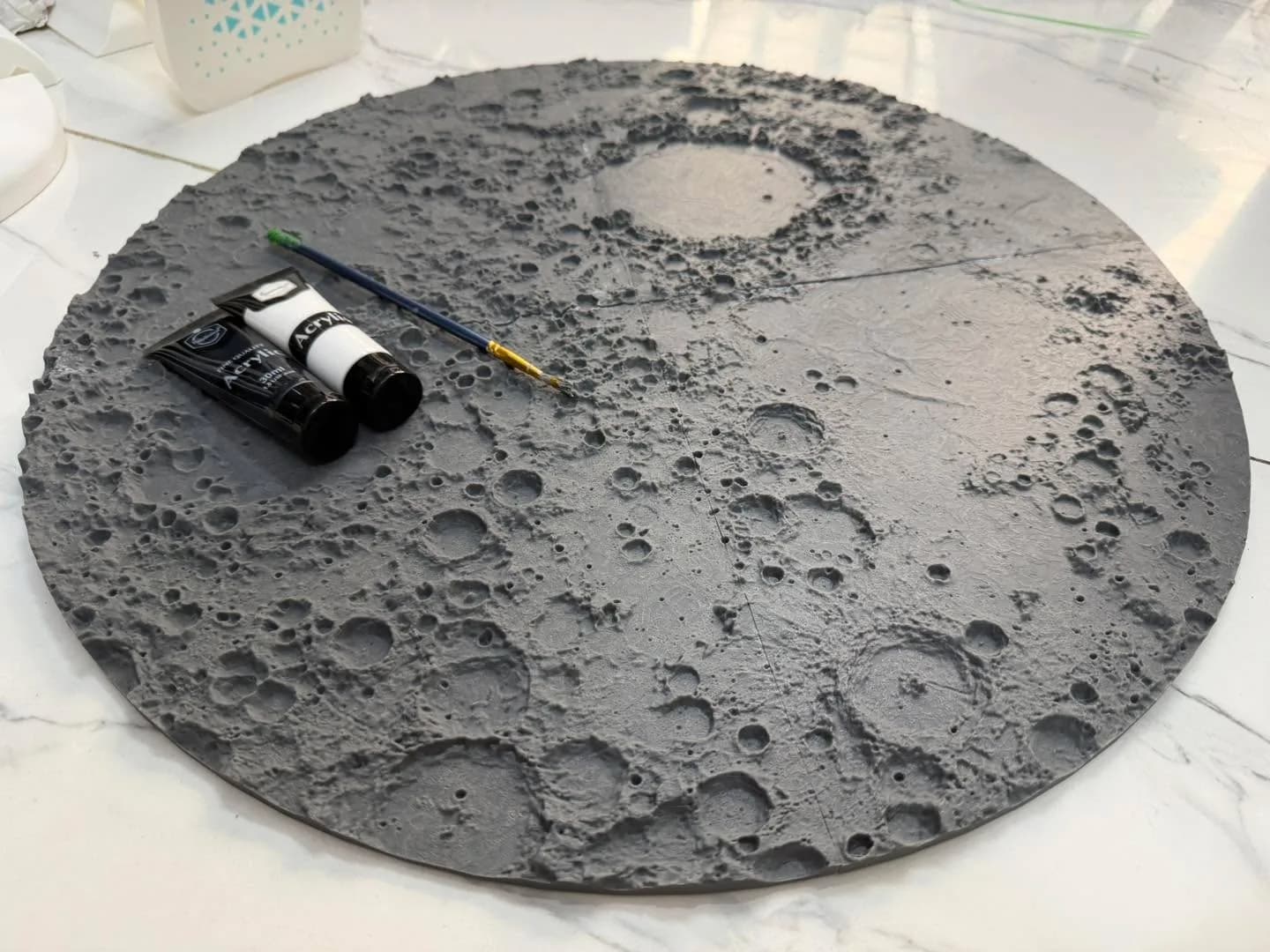 Grey 3D printed lunar relief disc before painting, with black and white acrylic paint tubes and a brush on top
