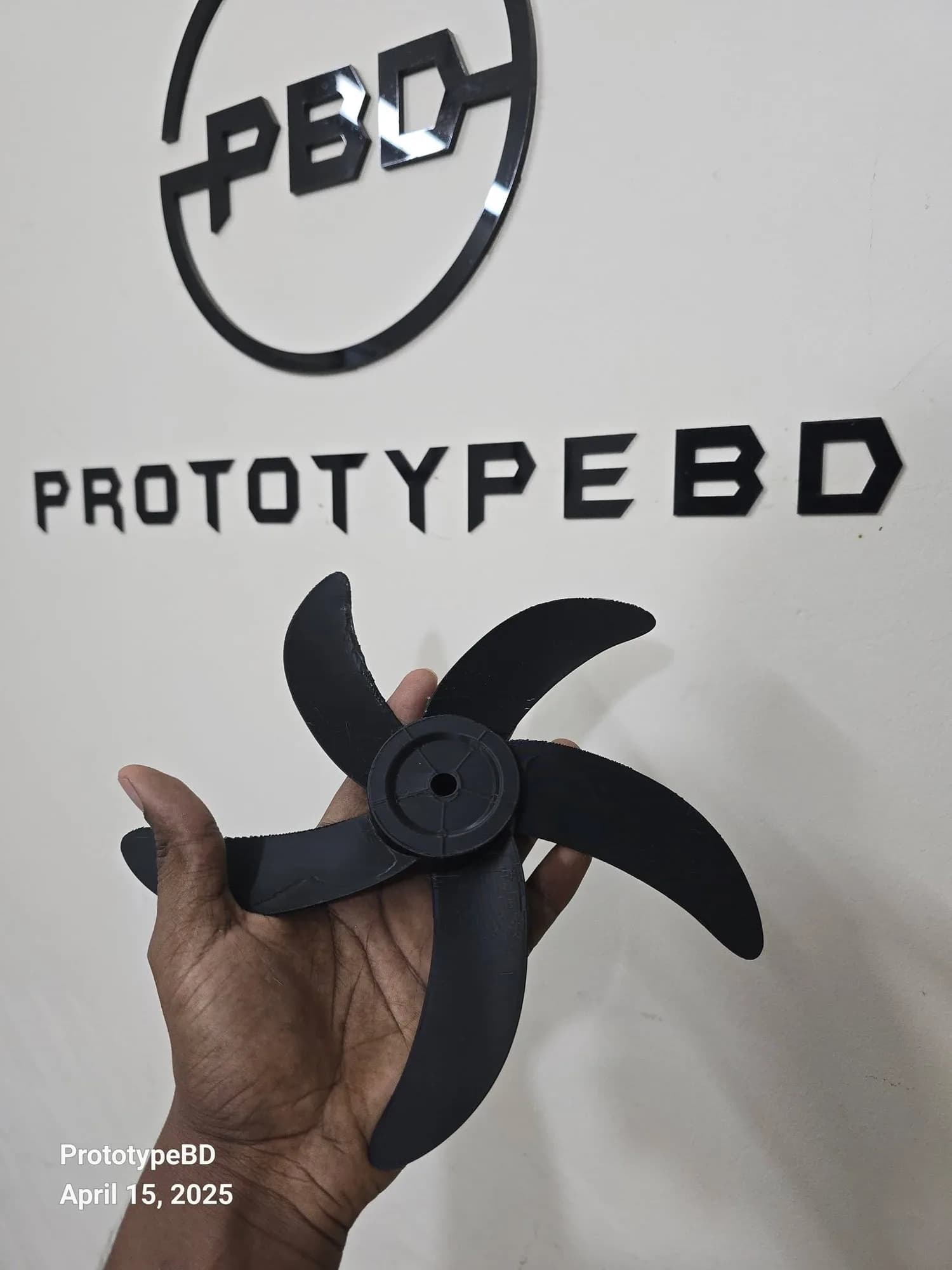 Hand holding the black 3D printed five-blade fan rotor in front of the PrototypeBD wall logo