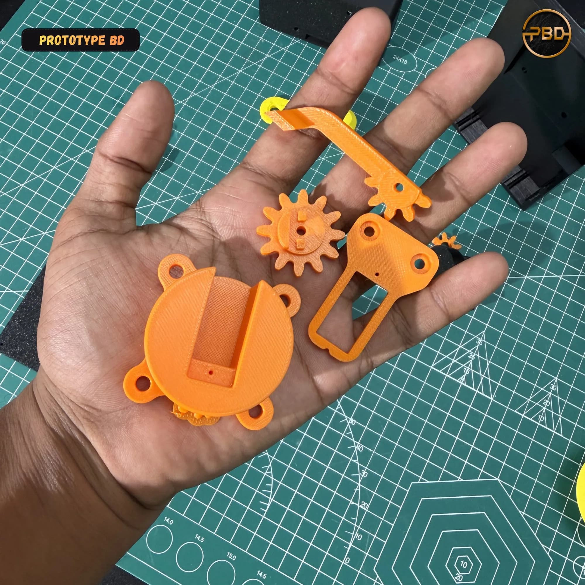 Hand holding orange 3D printed gripper parts: servo mount, spur gear, jaw and linkage arm