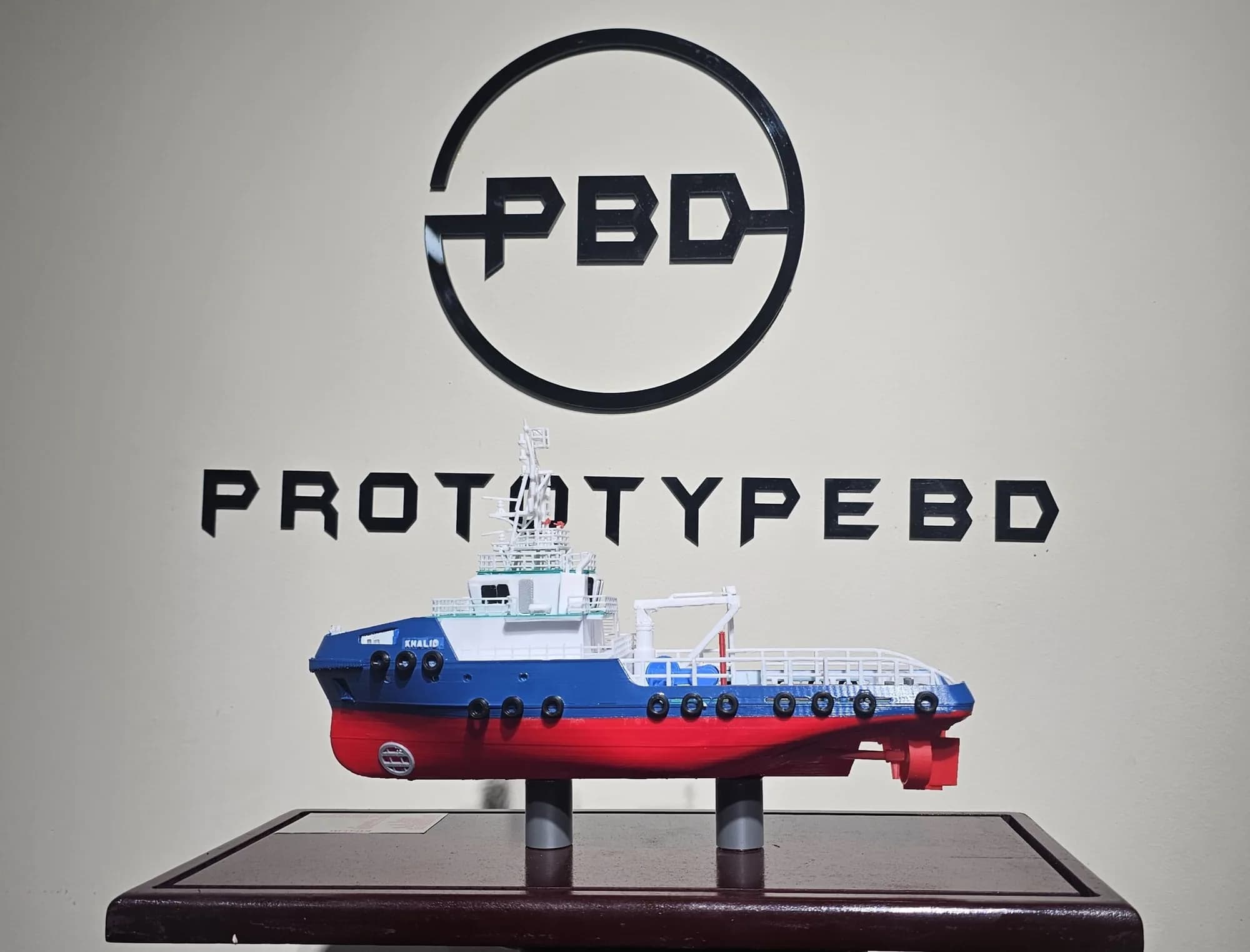Tugboat model in profile on its display base in front of the PrototypeBD wall logo