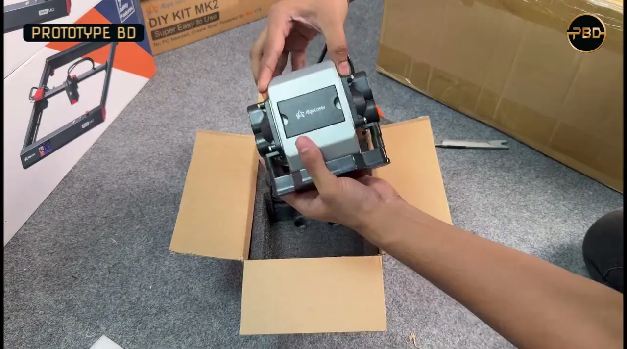 Hands lifting a grey AlgoLaser air pump out of its cardboard box on a carpeted floor