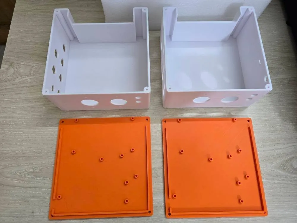 White 3D-printed aquaponics boxes with round cable holes and slot guides, beside orange lids with standoffs and screw holes