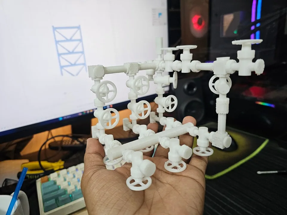 White 3D printed choke manifold model held over a desk, with valve handwheels and pipe header visible