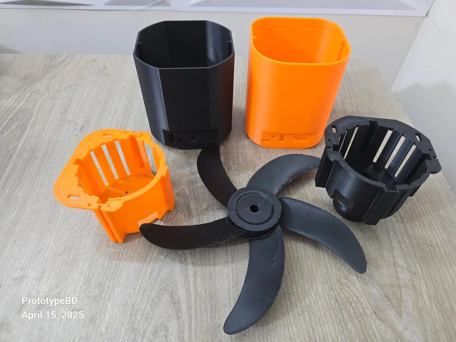3D printed fan parts on a table: four-blade black fan, orange and black motor housings and two body sections