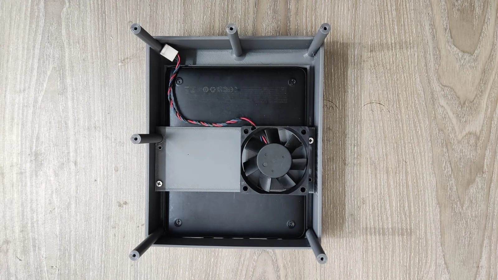 Grey 3D-printed enclosure tray from above, holding a black device, with a printed fan bracket, cooling fan and printed rods