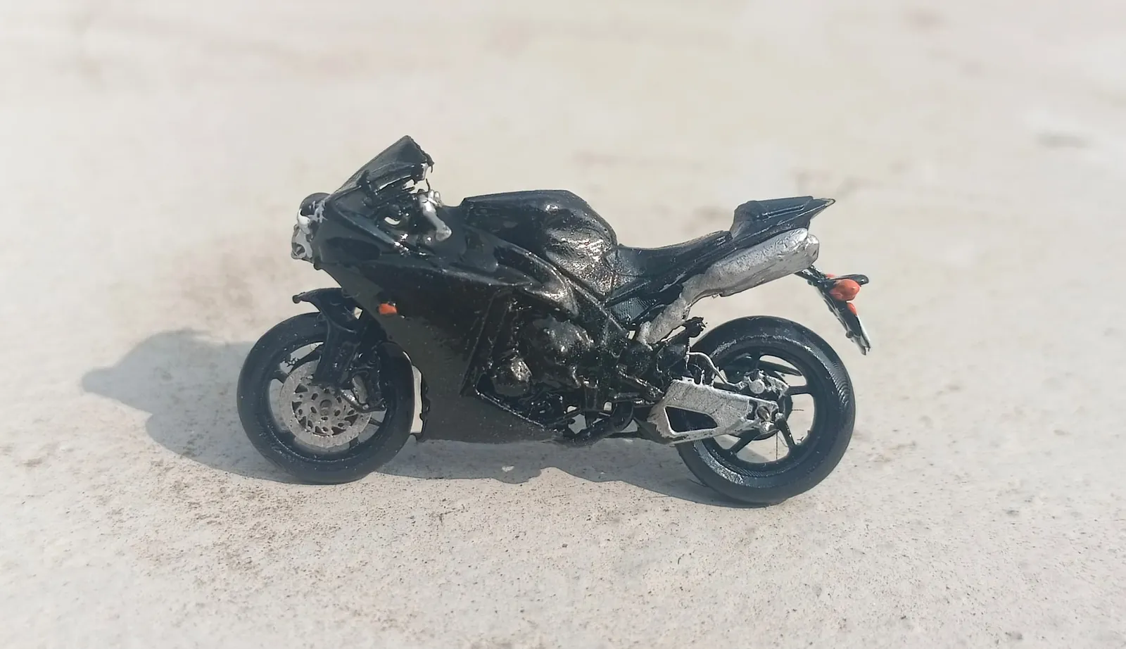Black resin 3D printed miniature sports motorbike model with silver exhaust and orange lights, on a concrete surface