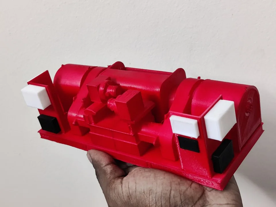 Red 3D printed oil-rig equipment model with a central cable drum and white and black details, held in one hand
