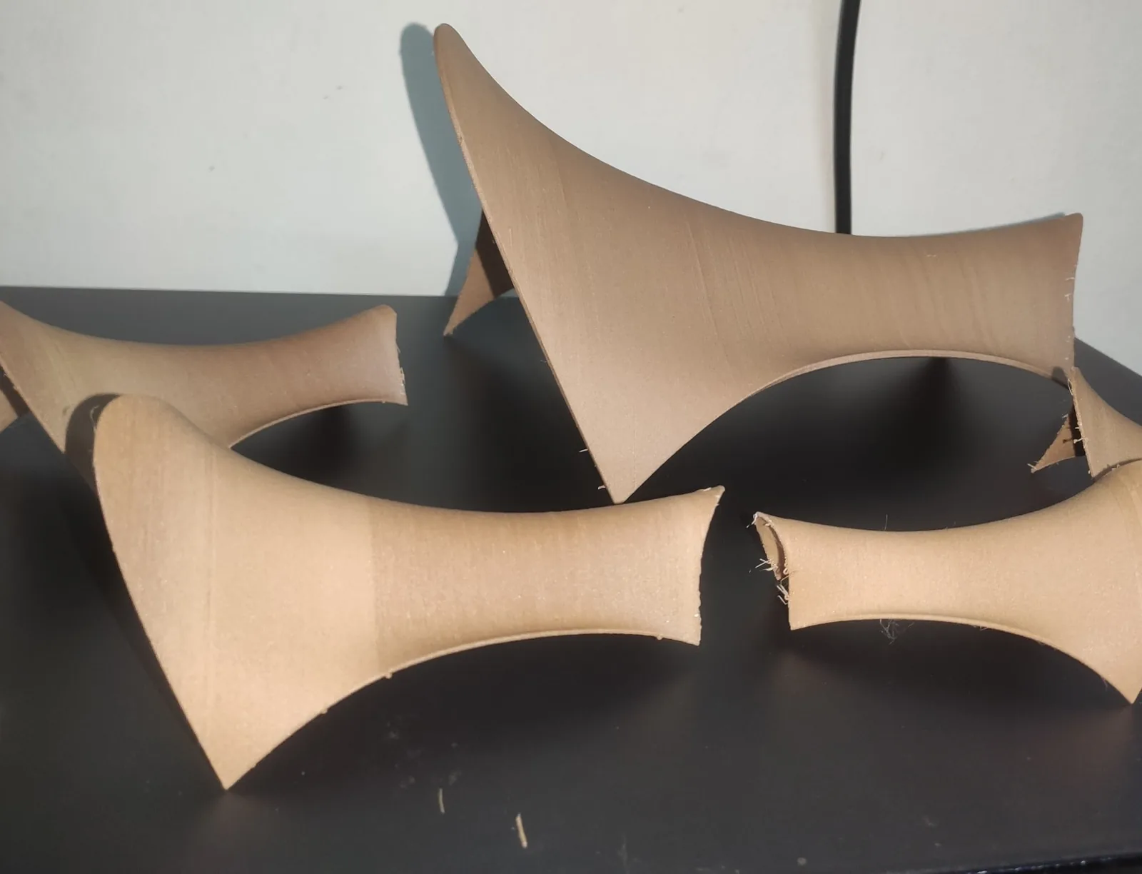 Five 3D-printed shell forms in light brown wood-filled filament with curved saddle shapes, on a dark table